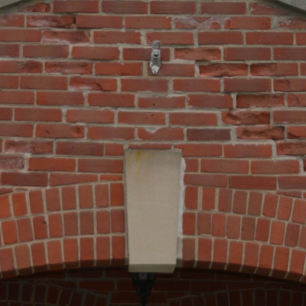 Red brick entryway with curve; a grey brick is placed at the top of the curve. There is a small metal hook-like item affixed to the brick above
